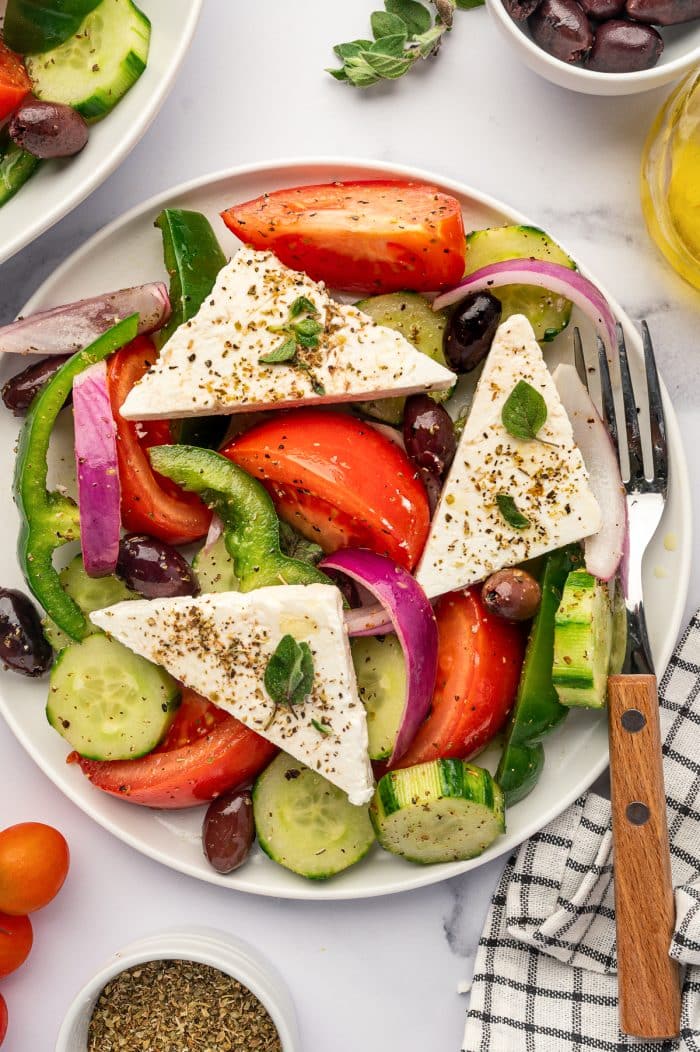 Close-up overhead view of traditional Greek salad with tomato wedges, cucumber rounds, red onion, green bell pepper, Kalamata olives, and triangular feta slabs sprinkled with dried oregano, with a wooden-handled fork and black-and-white checkered towel on the side.