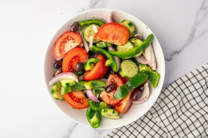 Overhead view of tossed Greek salad vegetables in a white bowl, including tomato wedges, cucumber rounds, sliced red onion, green bell pepper, Kalamata olives, dried oregano, salt, and black pepper beside a black-and-white checkered towel.