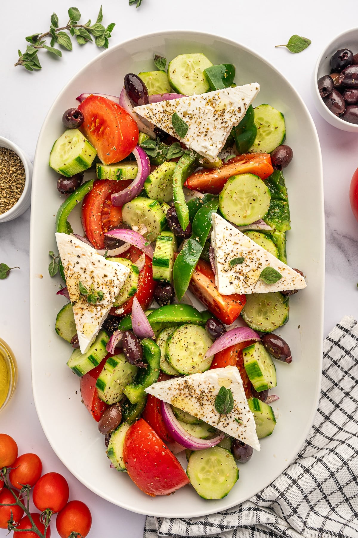 Overhead view of traditional Greek salad served on a light beige oval platter with tomato wedges, cucumber rounds, red onion, green bell pepper, Kalamata olives, and large triangular slabs of feta cheese topped with dried oregano and fresh oregano leaves, with extra olives, tomatoes, and a black-and-white checkered towel around the platter.