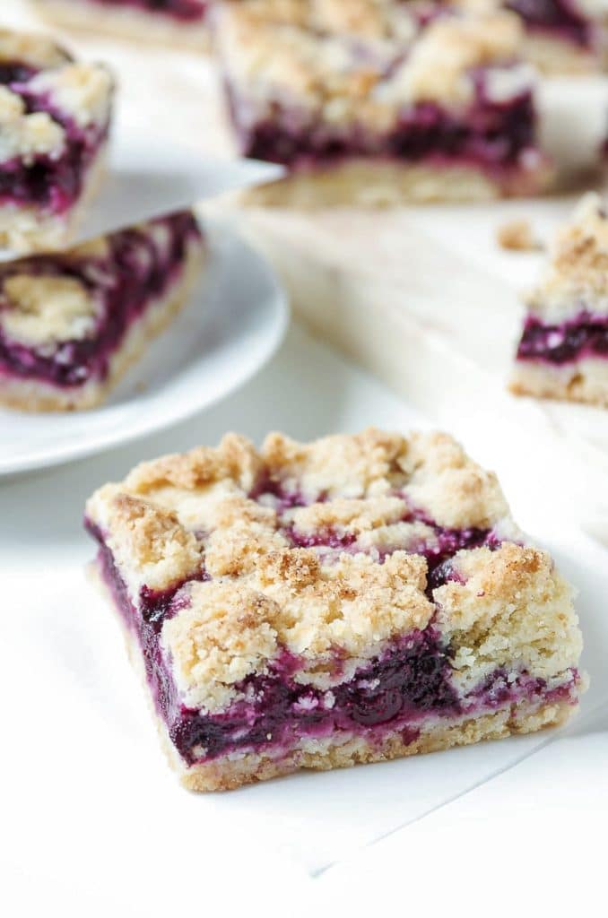 One blueberry bar with stacked blueberry bars in the background on a plate and cutting board