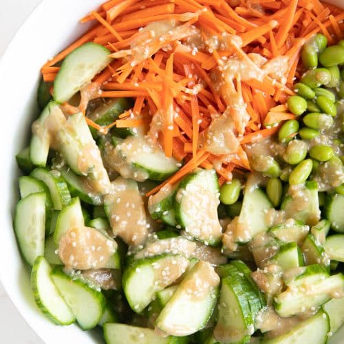 Large white salad bowl filled with julienned carrots, sliced cucumbers, and edamame and drizzled with homemade miso dressing.