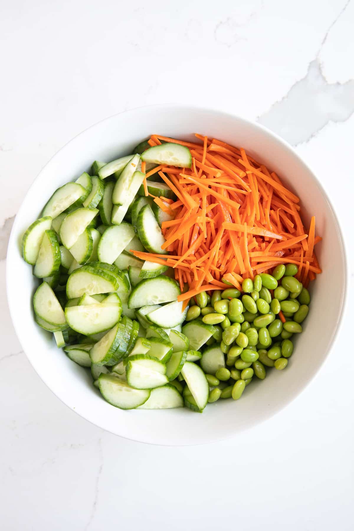 White salad bowl filled with julienned carrots, edamame, and sliced cucumbers.