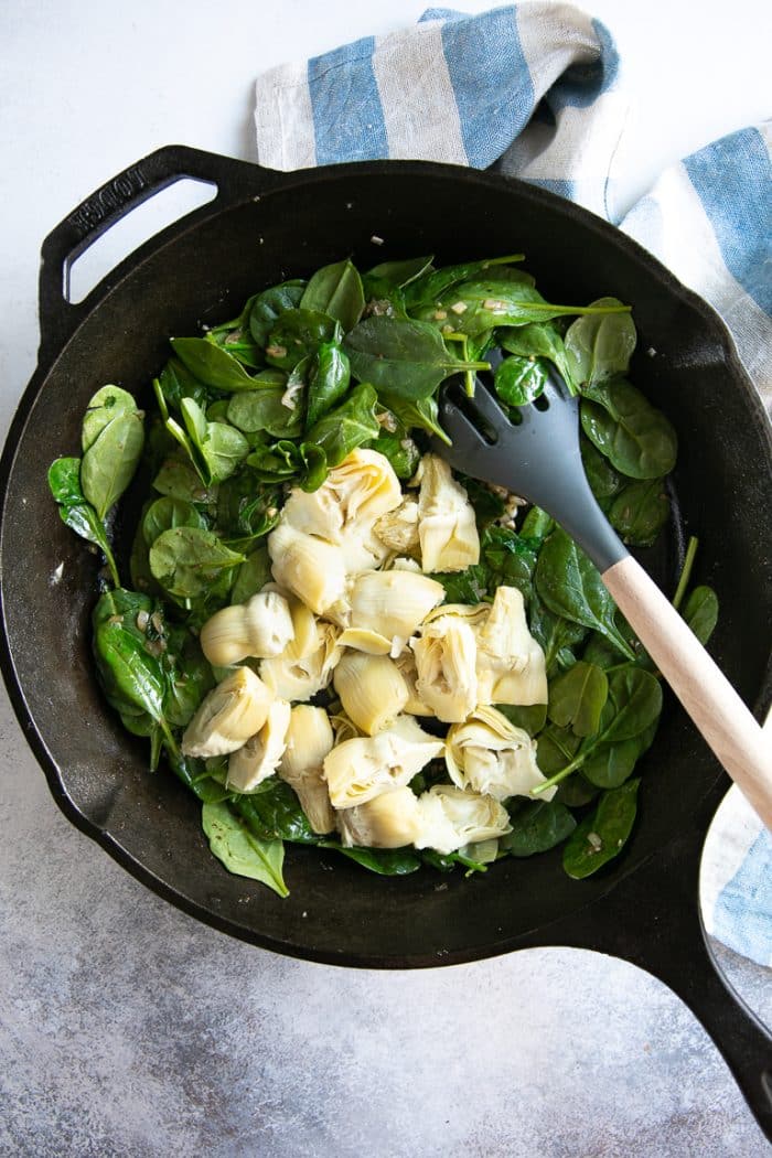 Overhead image of a large cast iron skillet filled with fresh spinach and artichoke hearts.