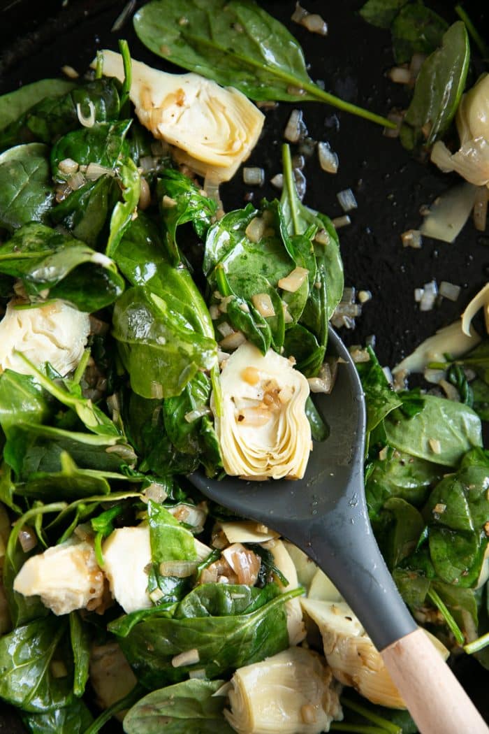 Image of a cast iron skillet filled with artichoke hearts and spinach cooking in a large cast iron skillet with shallots and garlic.
