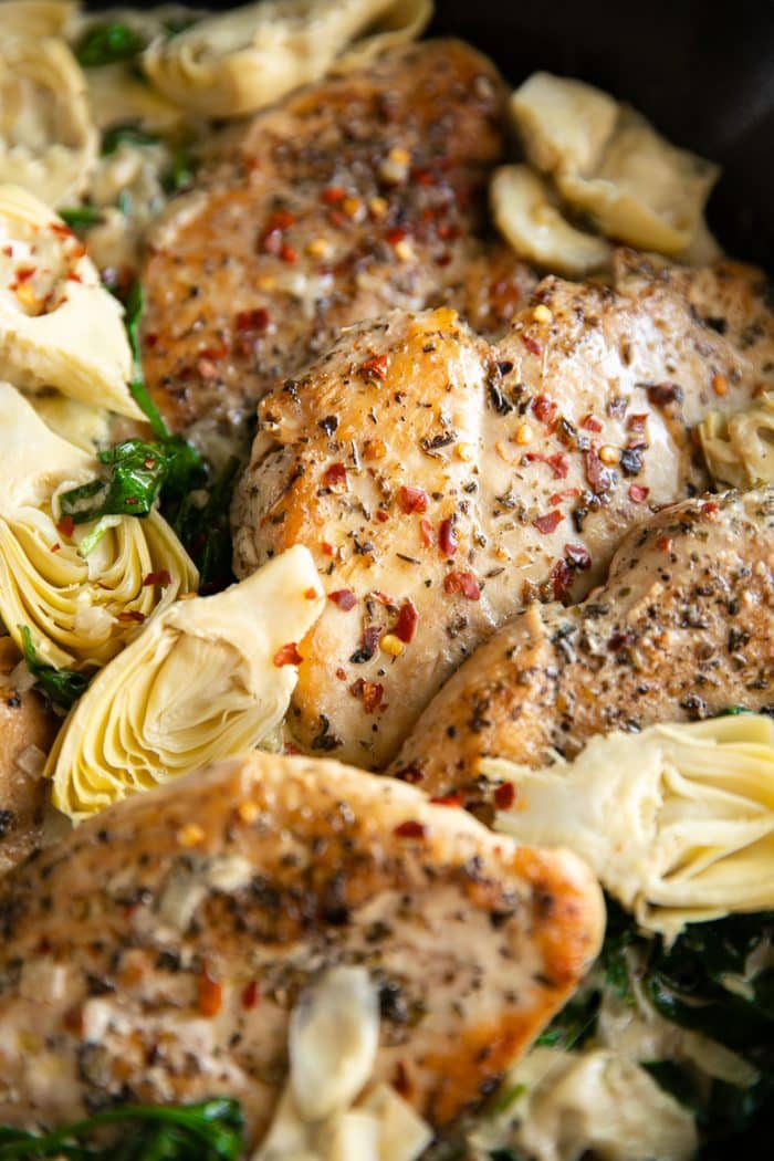 Image of cooked chicken breasts surrounded by artichoke hearts and spinach in a light cream sauce all in a cast-iron skillet.