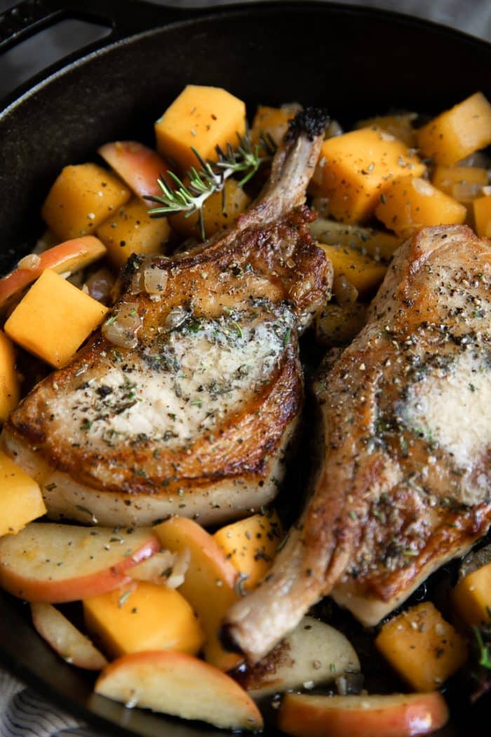 Close-up image of two bone-in pork chops cooked with apples and butternut squash and topped with butter and herbs