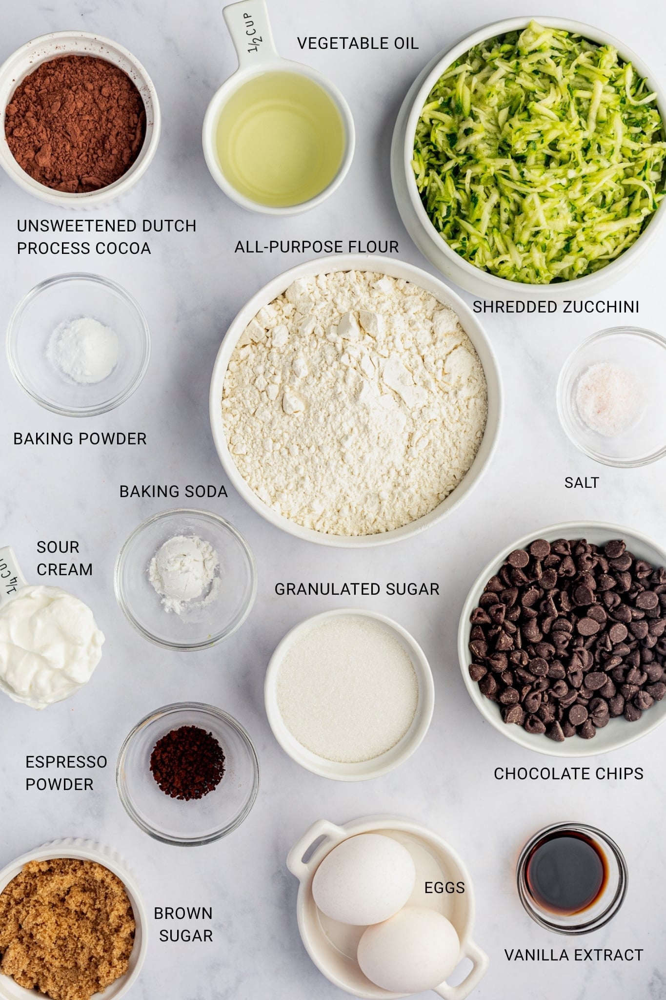 Overhead view of the ingredients for double chocolate zucchini muffins arranged on a white marble surface in white and pale green ceramic bowls, clear glass prep bowls, and measuring cups. The image includes cocoa powder, pale yellow vegetable oil, bright green shredded zucchini, all-purpose flour, baking powder, baking soda, pink-tinged salt, sour cream, granulated sugar, semisweet chocolate chips, espresso powder, brown sugar, two white eggs, and dark vanilla extract. Black text overlays label each ingredient: "UNSWEETENED DUTCH PROCESS COCOA," "VEGETABLE OIL," "SHREDDED ZUCCHINI," "ALL-PURPOSE FLOUR," "BAKING POWDER," "BAKING SODA," "SALT," "SOUR CREAM," "GRANULATED SUGAR," "CHOCOLATE CHIPS," "ESPRESSO POWDER," "BROWN SUGAR," "EGGS," and "VANILLA EXTRACT." There are no garnishes.