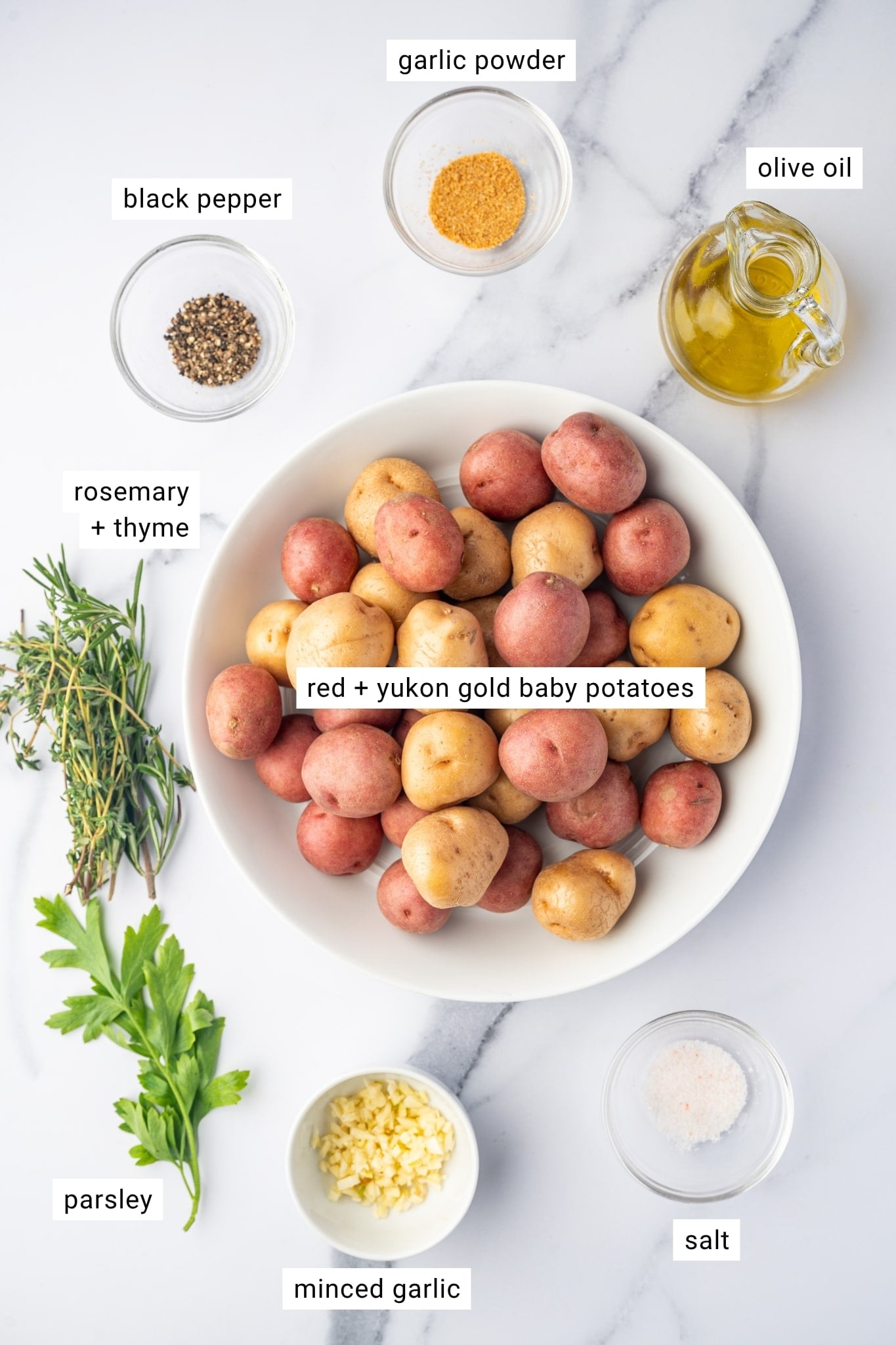 Overhead view of the ingredients for garlic roasted potatoes arranged on a white marble surface. A large white bowl in the center holds red and Yukon Gold baby potatoes, surrounded by fresh rosemary and thyme sprigs, fresh parsley, a small white bowl of minced garlic, a clear bowl of salt, a clear bowl of cracked black pepper, a clear bowl of garlic powder, and a glass cruet filled with golden olive oil. White text labels identify "red + yukon gold baby potatoes," "rosemary + thyme," "parsley," "minced garlic," "salt," "black pepper," "garlic powder," and "olive oil."