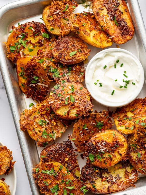 Overhead view of crispy Parmesan potatoes arranged on a rectangular silver serving tray, garnished generously with chopped green parsley and grated Parmesan, with a small white bowl of creamy sauce nestled among the golden-brown potatoes.