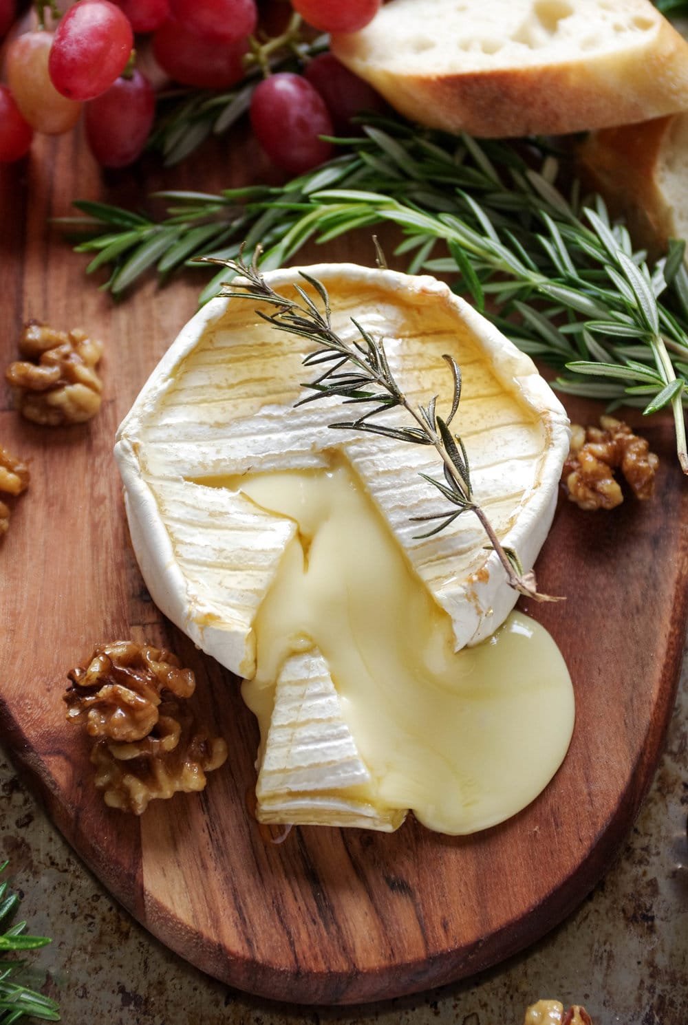 Gooey baked brie with maple candied walnuts on a small wood serving board with grapes and sliced bread.