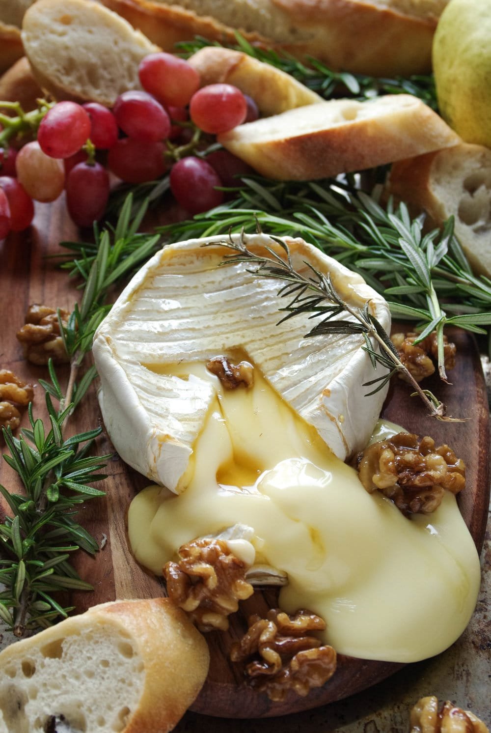 Gooey baked brie with maple candied walnuts on a small wood serving board with grapes and sliced bread.