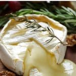 A wheel of baked brie cheese with a wedge cut out, allowing the gooey, melted cheese to flow onto a wooden serving board. The brie is drizzled with honey, garnished with a sprig of fresh rosemary, and surrounded by candied walnuts.