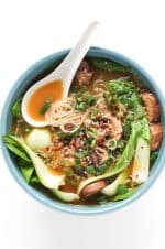 Ginger garlic noodle soup with bok choy, mushrooms, rice noodles, green onions, sesame seeds, and chili flakes in a blue bowl.