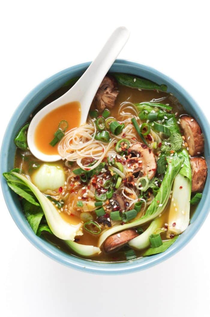 Ginger garlic noodle soup with bok choy, mushrooms, rice noodles, green onions, sesame seeds, and chili flakes in a blue bowl.