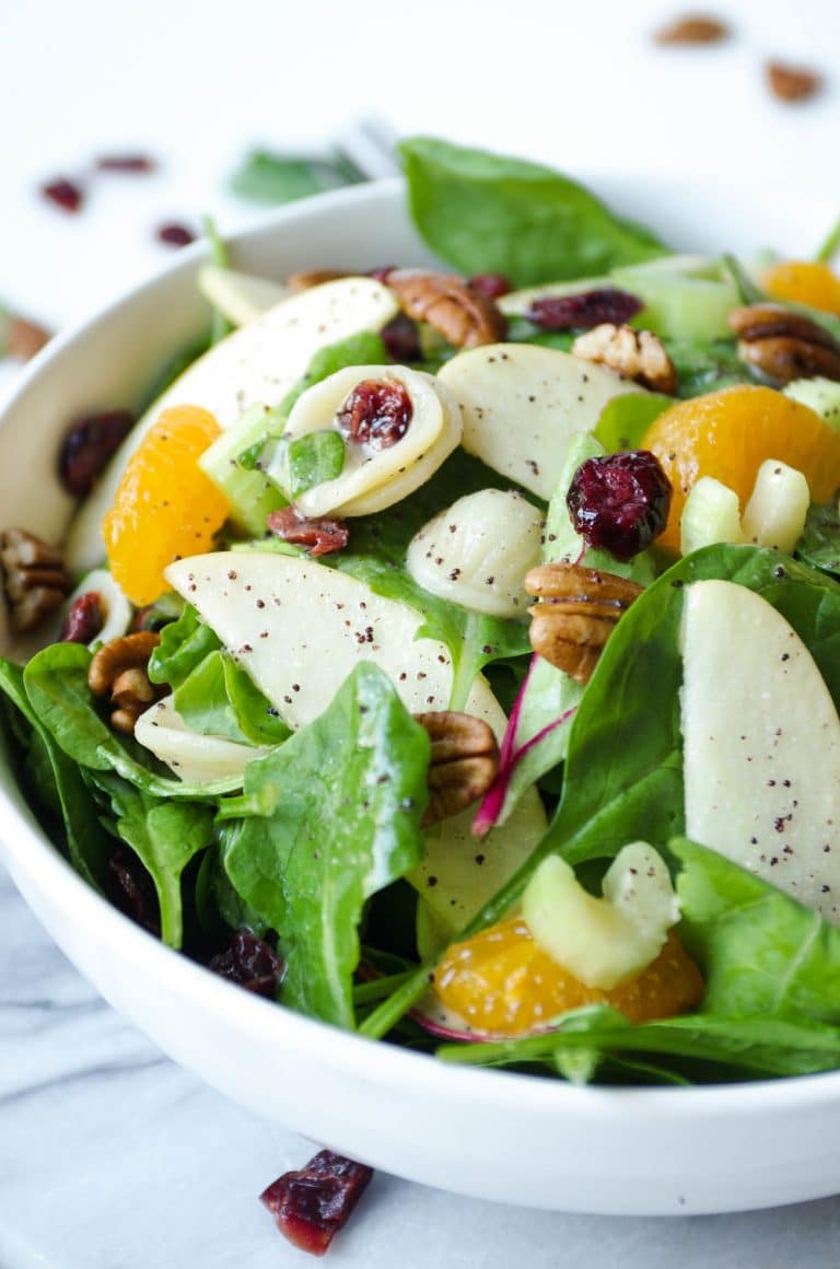 Close up image of a large serving bowl filled with a light spinach pasta salad filled with mixed greens, cooked pasta, mandarin orange slices, dried cranberries, whole pecans, and poppyseed dressing.