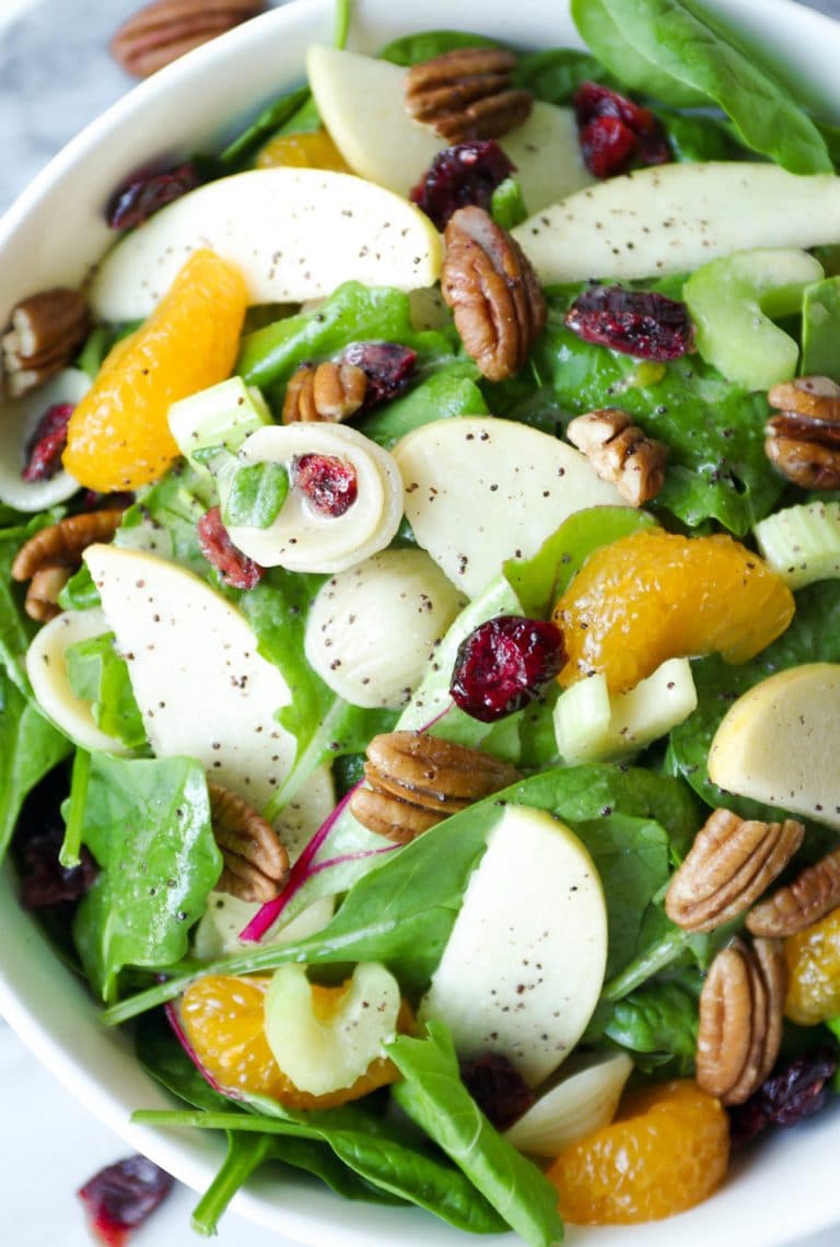 Close up image of a large serving bowl filled with a light spinach pasta salad filled with mixed greens, cooked pasta, mandarin orange slices, dried cranberries, whole pecans, and poppyseed dressing.