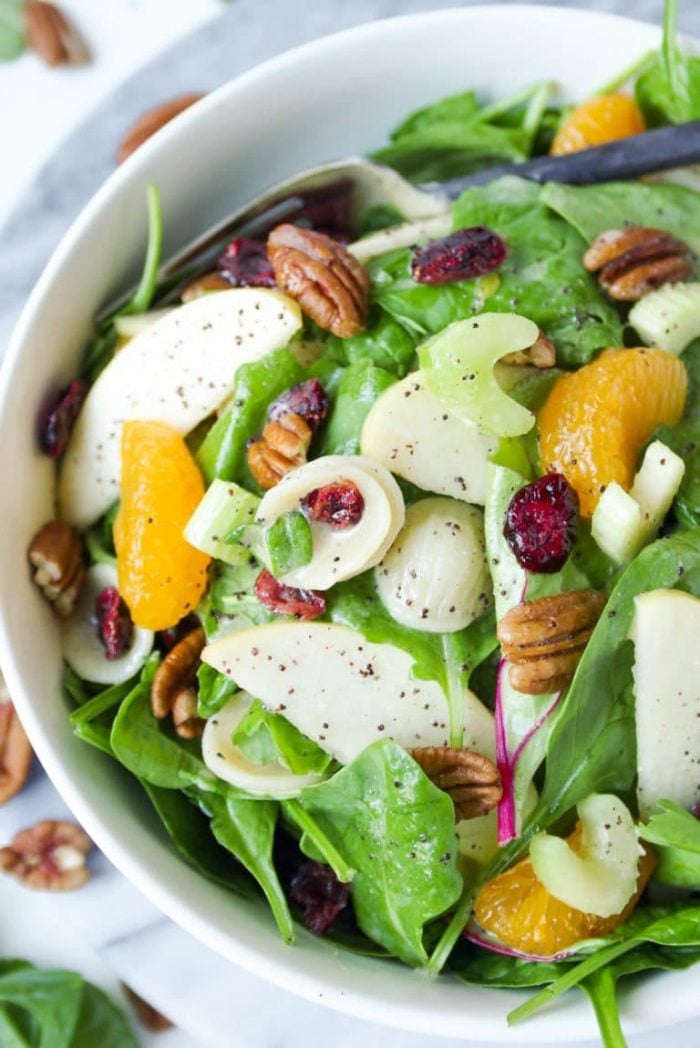 Close up image of a large serving bowl filled with a light spinach pasta salad filled with mixed greens, cooked pasta, mandarin orange slices, dried cranberries, whole pecans, and poppyseed dressing.