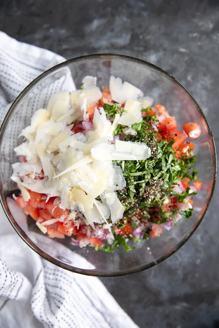A glass bowl filled with chopped tomatoes, onions, salt, pepper, garlic, fresh basic, and shaved parmesan cheese.