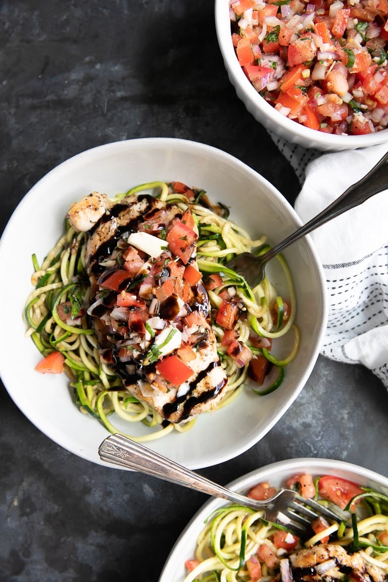 White bowls filled with zucchini noodles and bruschetta chicken topped with balsamic glaze.