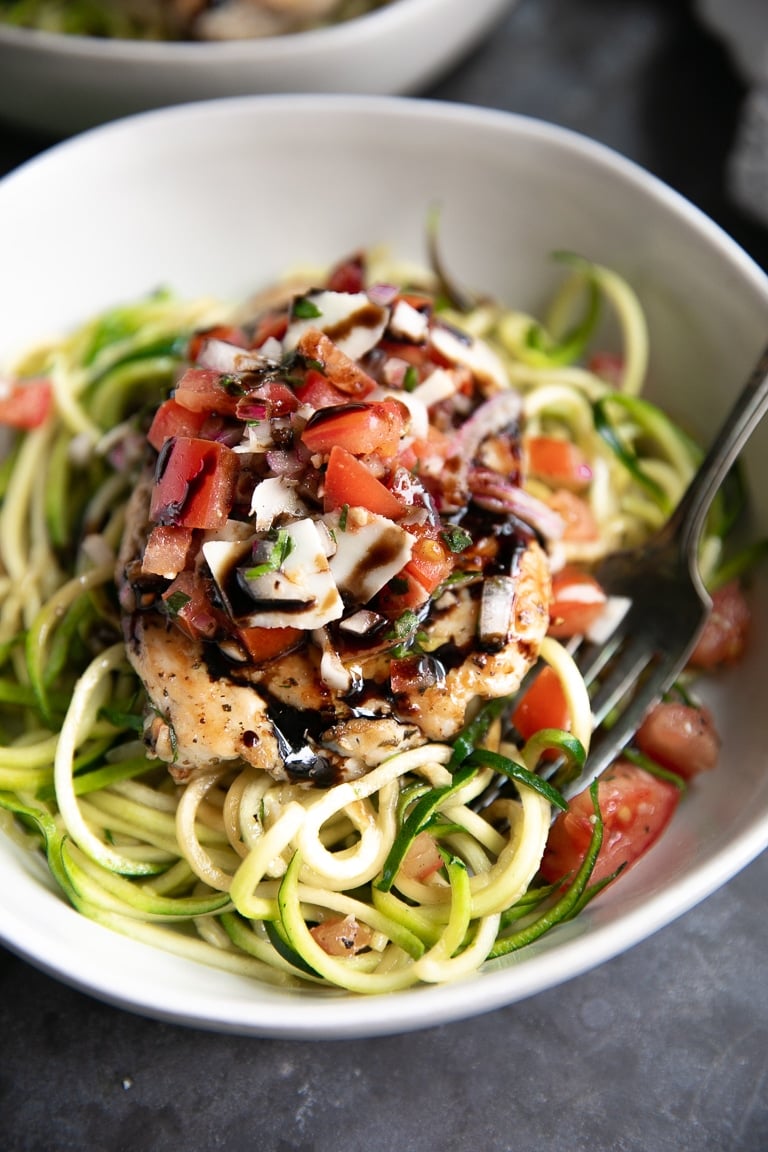 Serving bowl filled with cooked zucchini noodles and topped with a cooked chicken breast topped with tomato bruschetta topping.