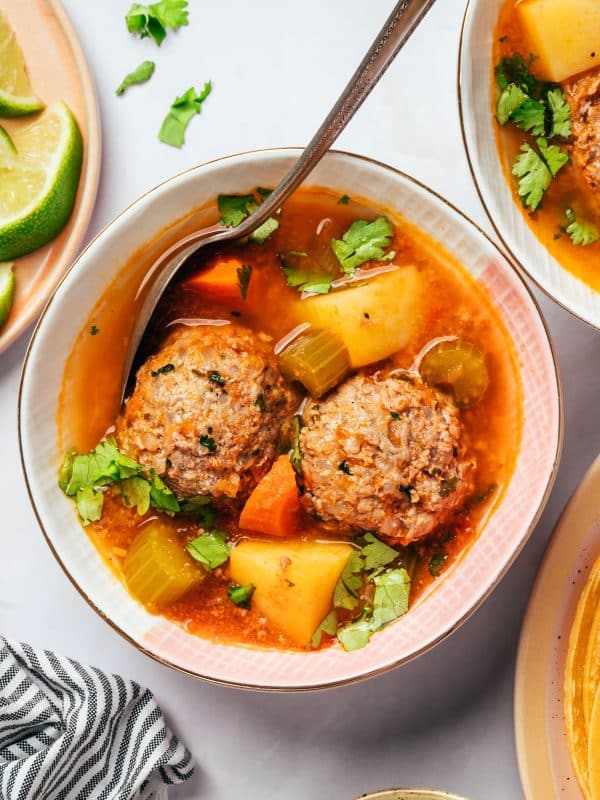 A white bowl of albondigas soup containing two large cooked meatballs, cubed potatoes, orange carrots, celery, and scattered cilantro in a tomato broth. A silver spoon rests inside the bowl, with lime wedges and a second serving of soup nearby.