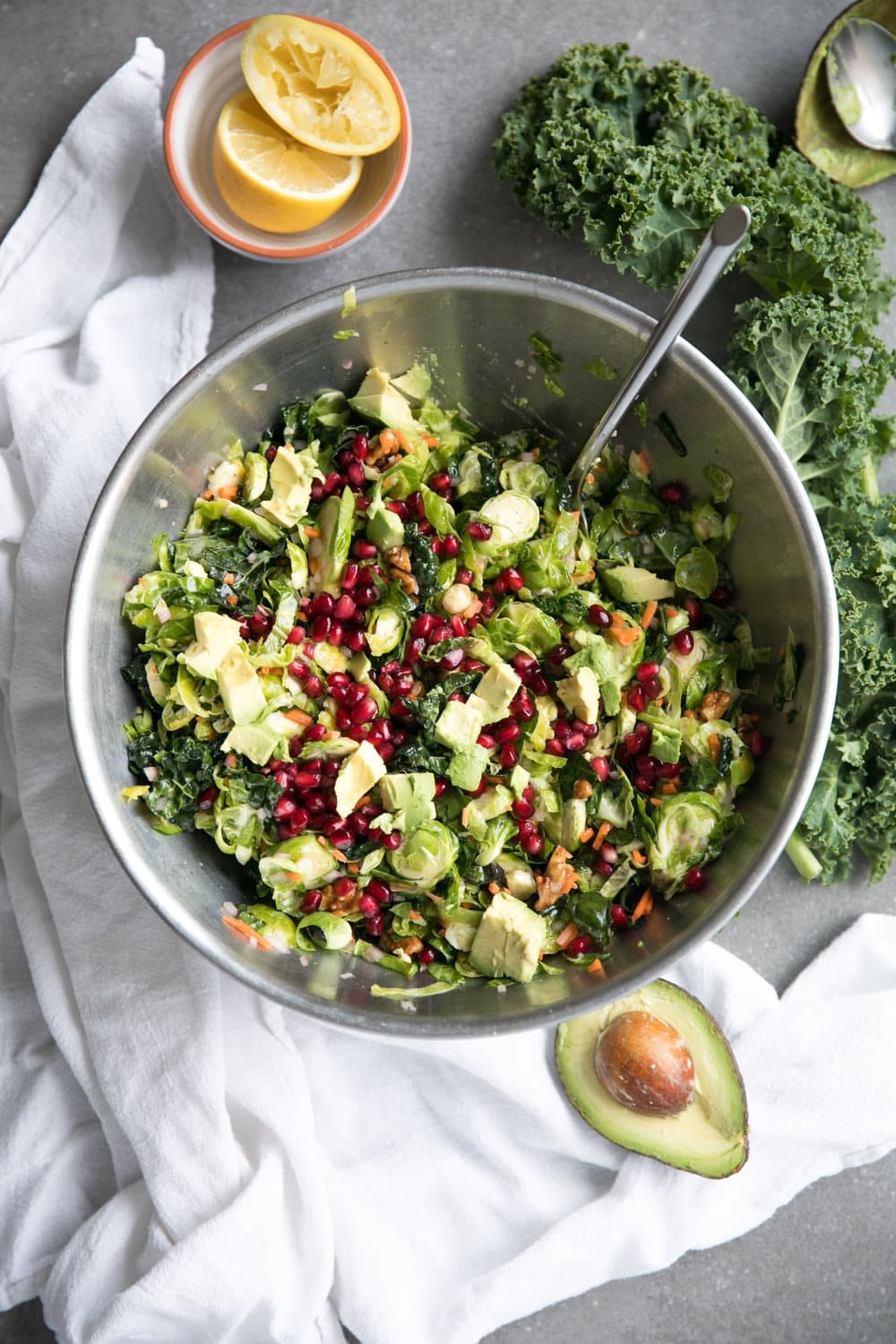 Overhead image of a mixing bowl filled with shredded Brussels sprouts, kale, avocado, grated carrot, pomegranate arils, tossed in a light lemon vinaigrette.