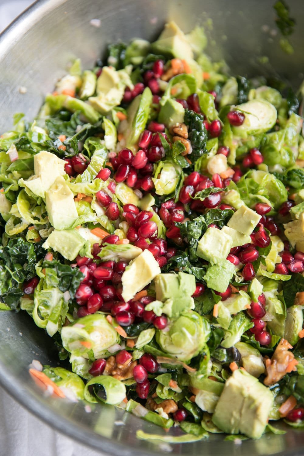 Close up of a large mixing bowl filled with shredded Brussels sprouts, kale, avocado, grated carrot, pomegranate arils, tossed in a light lemon vinaigrette.