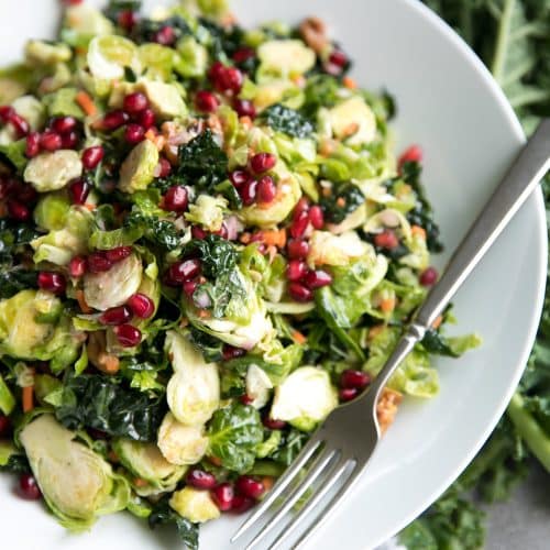 Close up of a large white salad bowl filled with shredded Brussels sprouts, kale, avocado, grated carrot, pomegranate arils, tossed in a light lemon vinaigrette.
