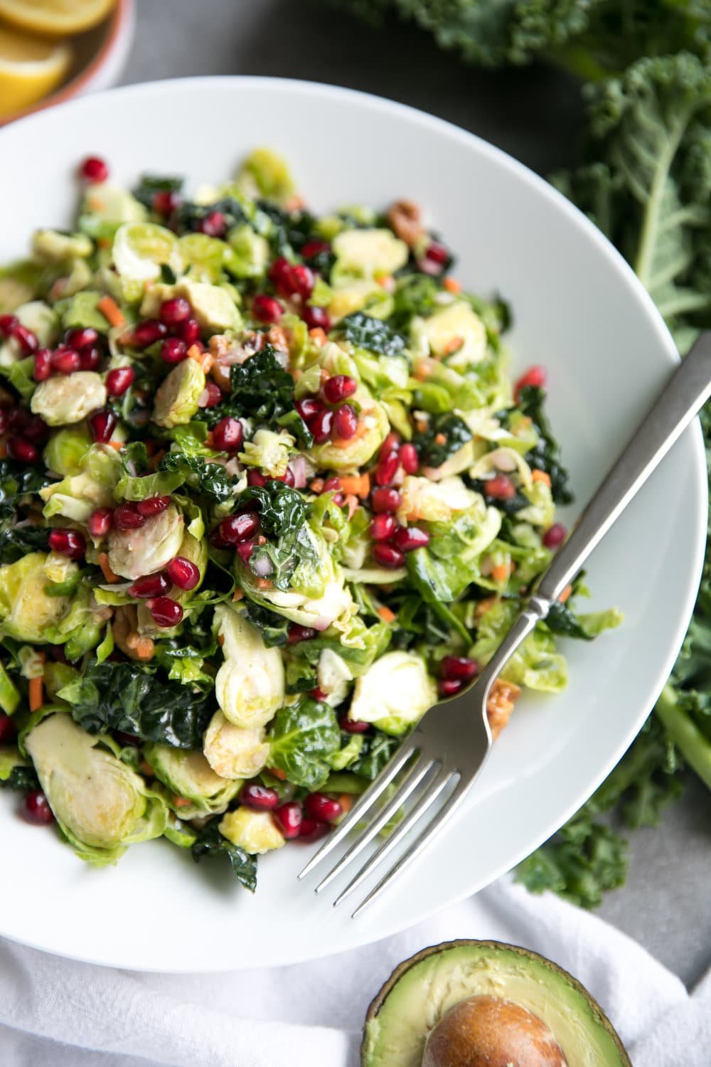 Close up of a large white salad bowl filled with shredded Brussels sprouts, kale, avocado, grated carrot, pomegranate arils, tossed in a light lemon vinaigrette.