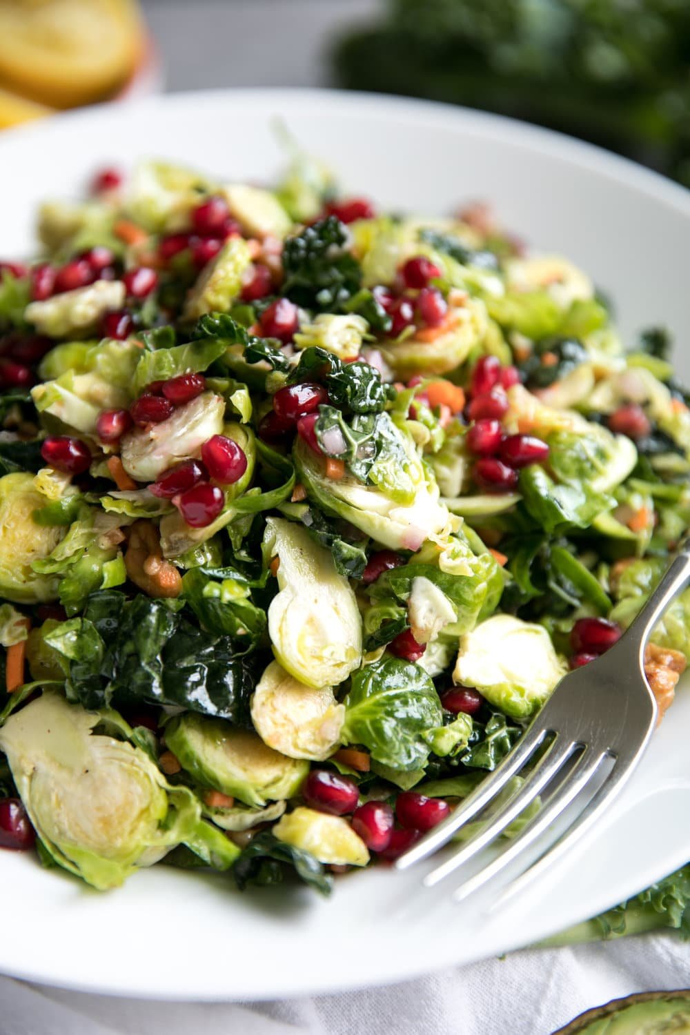 Close up of a large white salad bowl filled with shredded Brussels sprouts, kale, avocado, grated carrot, pomegranate arils, tossed in a light lemon vinaigrette.