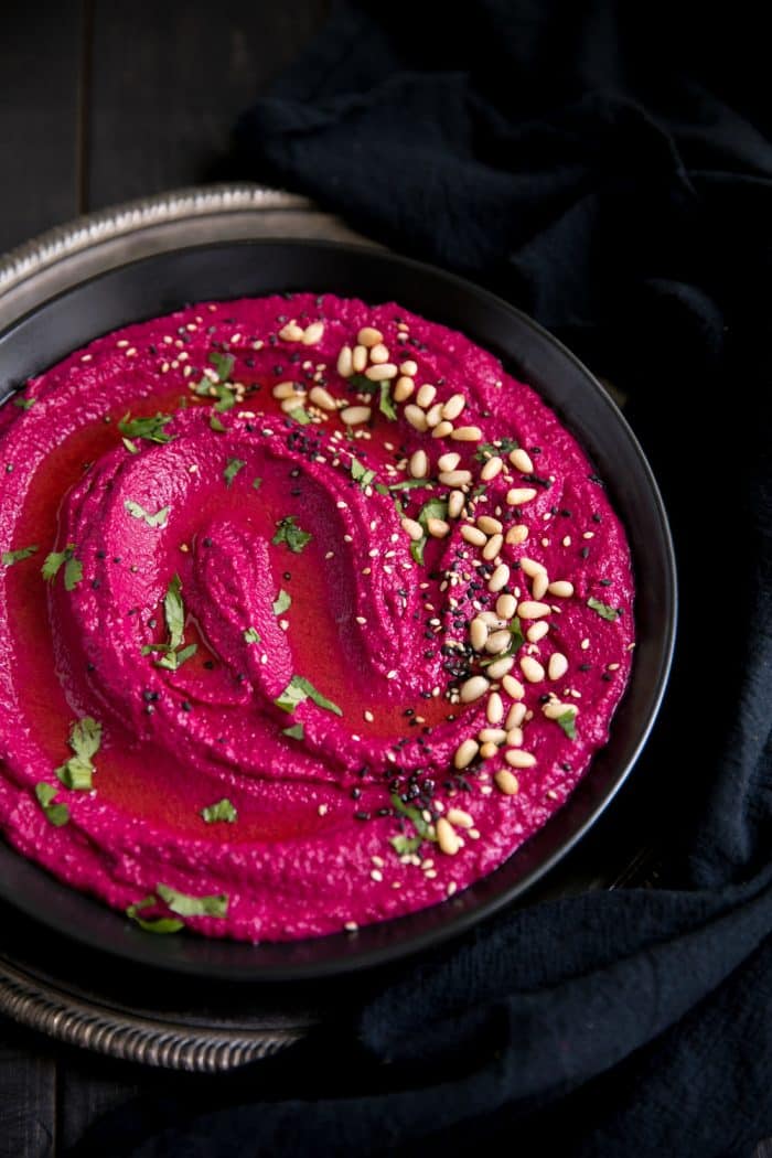 Large black serving bowl filled with bright pink beet hummus garnished with olive oil, pine nuts, and chopped parsley.