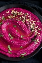 Large black serving bowl filled with bright pink beet hummus garnished with olive oil, pine nuts, and chopped parsley.