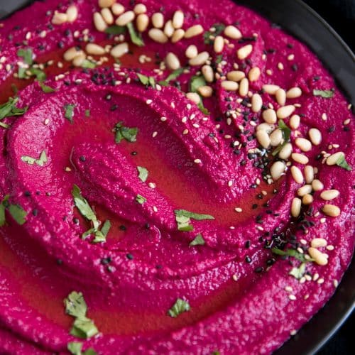 Large black serving bowl filled with bright pink beet hummus garnished with olive oil, pine nuts, and chopped parsley.