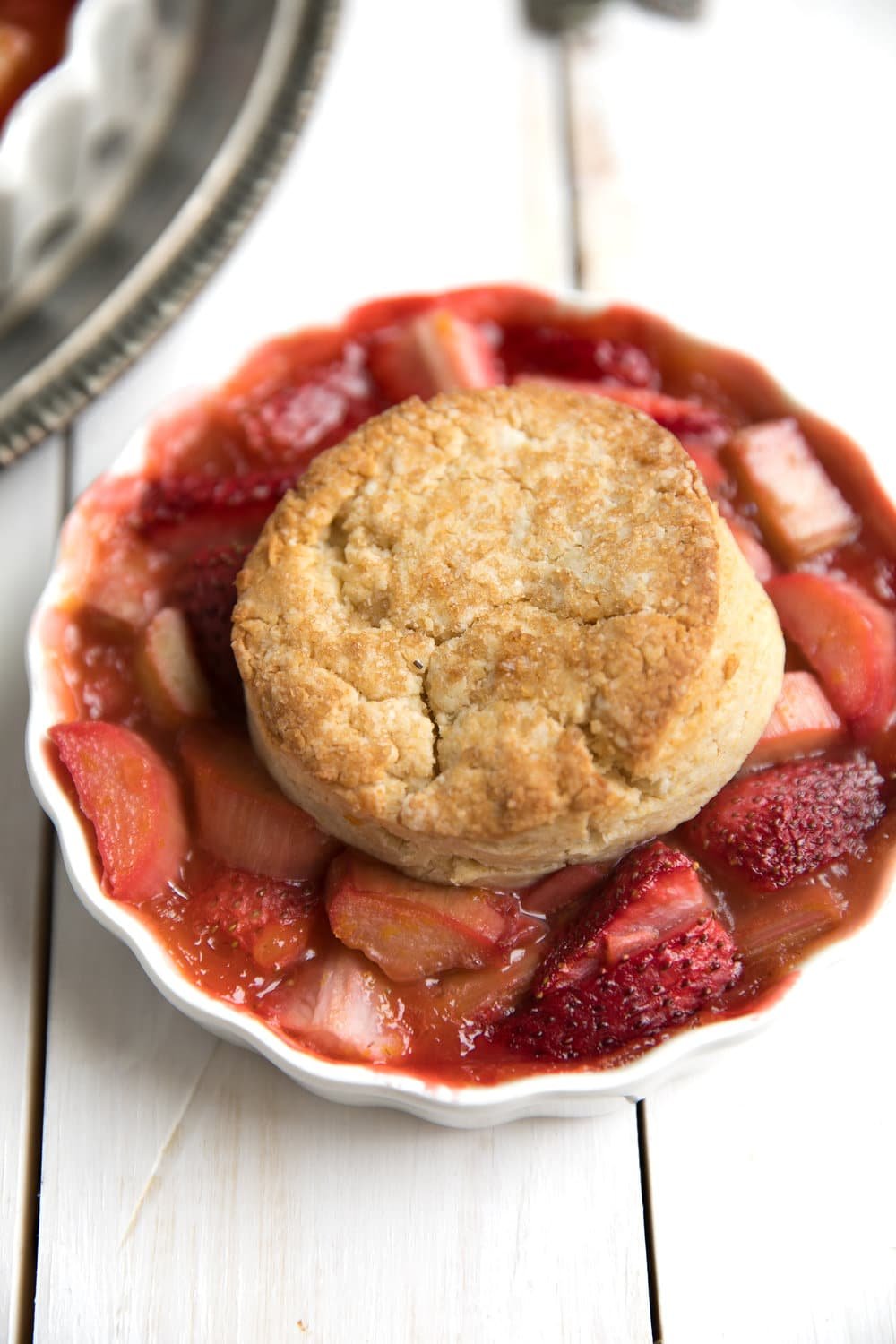 Single serving of Strawberry Rhubarb Cobbler on a white table with ripe strawberries that have black seeds