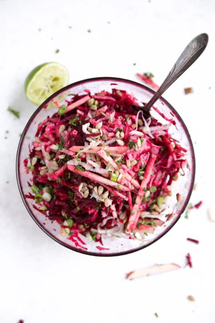 Glass bowl on a white table filled with raw beets, shredded kohlrabi, apples, green onion, feta, and sunflower seeds all mixed together.