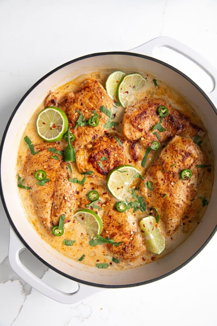 Large white enameled skillet filled with thinly sliced chicken breasts simmering in a homemade sauce made with coconut milk, chicken broth, and lime juice, and garnished with lime slices and fresh cilantro.