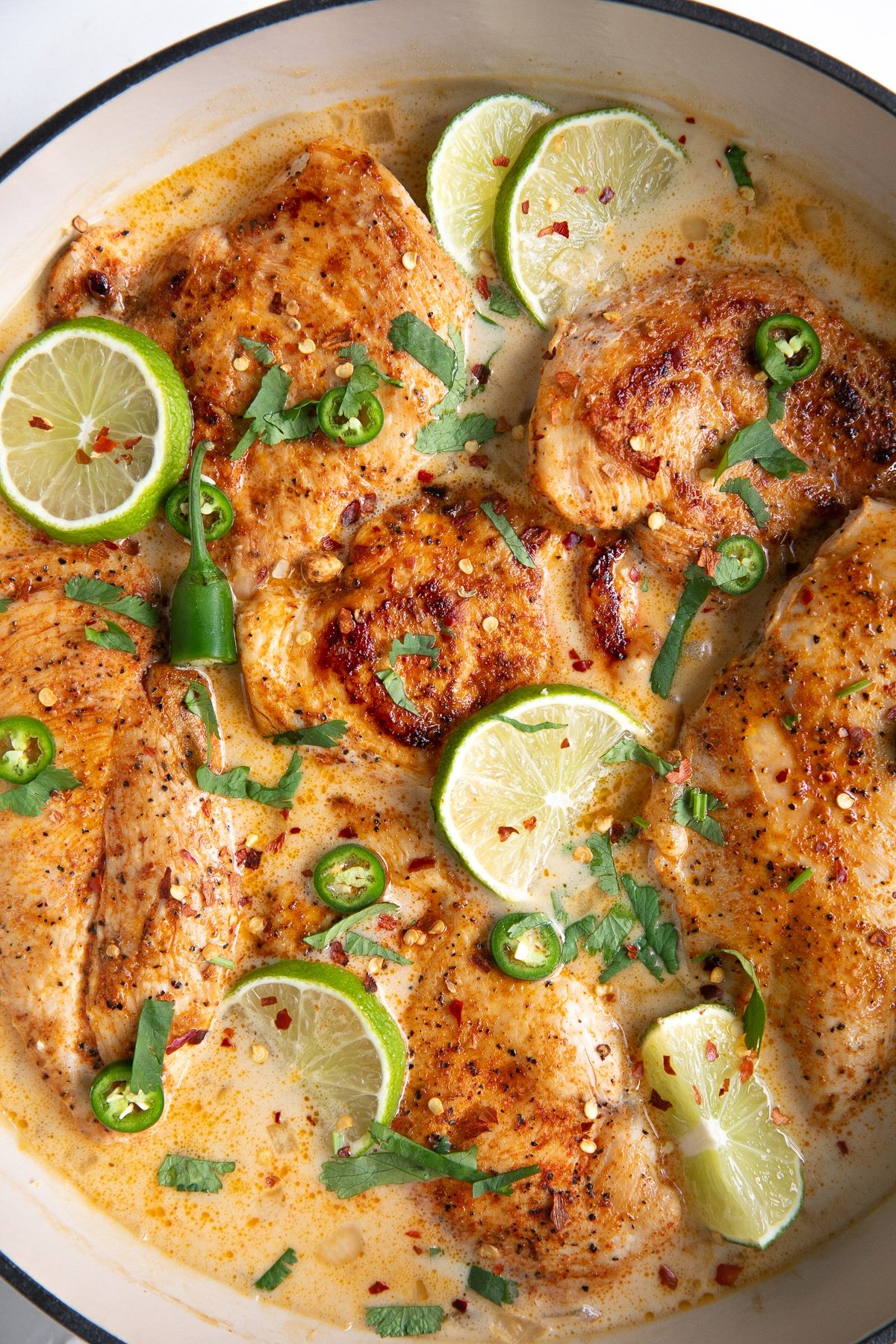Large white enameled skillet filled with thinly sliced chicken breasts simmering in a homemade sauce made with coconut milk, chicken broth, and lime juice, and garnished with lime slices and fresh cilantro.