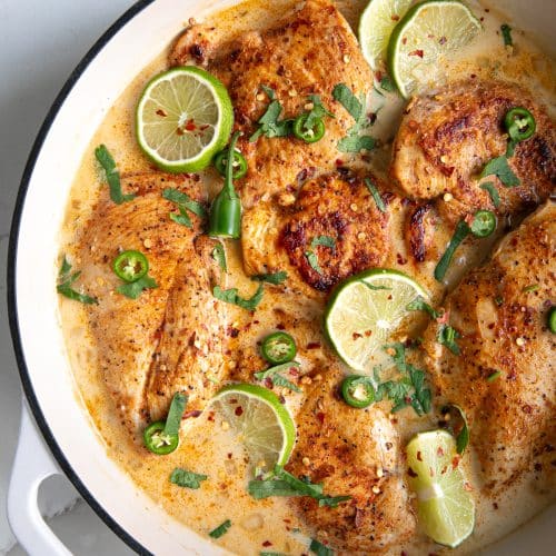 Large white enameled skillet filled with thinly sliced chicken breasts simmering in a homemade sauce made with coconut milk, chicken broth, and lime juice, and garnished with lime slices and fresh cilantro.