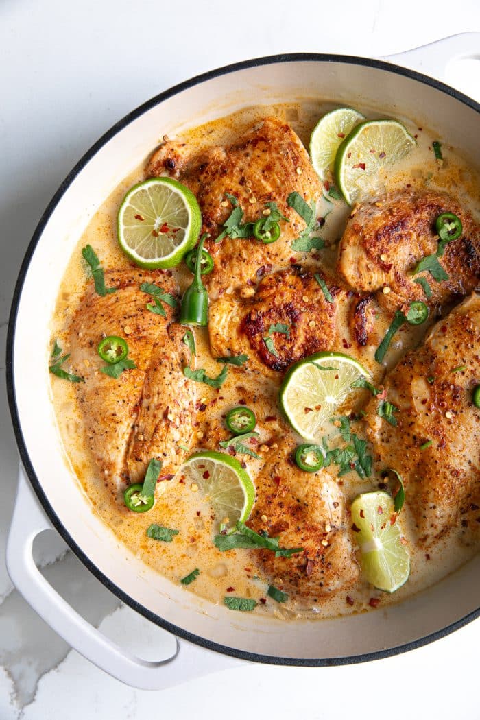 Large enamelled white pan filled with thinly sliced chicken breasts that simmer in a homemade sauce made with coconut milk, chicken broth and lemon juice, and garnished with lime slices and fresh cilantro.