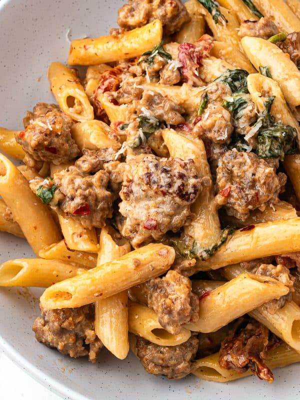 Creamy sausage pasta served in a white speckled bowl with a wooden spoon.