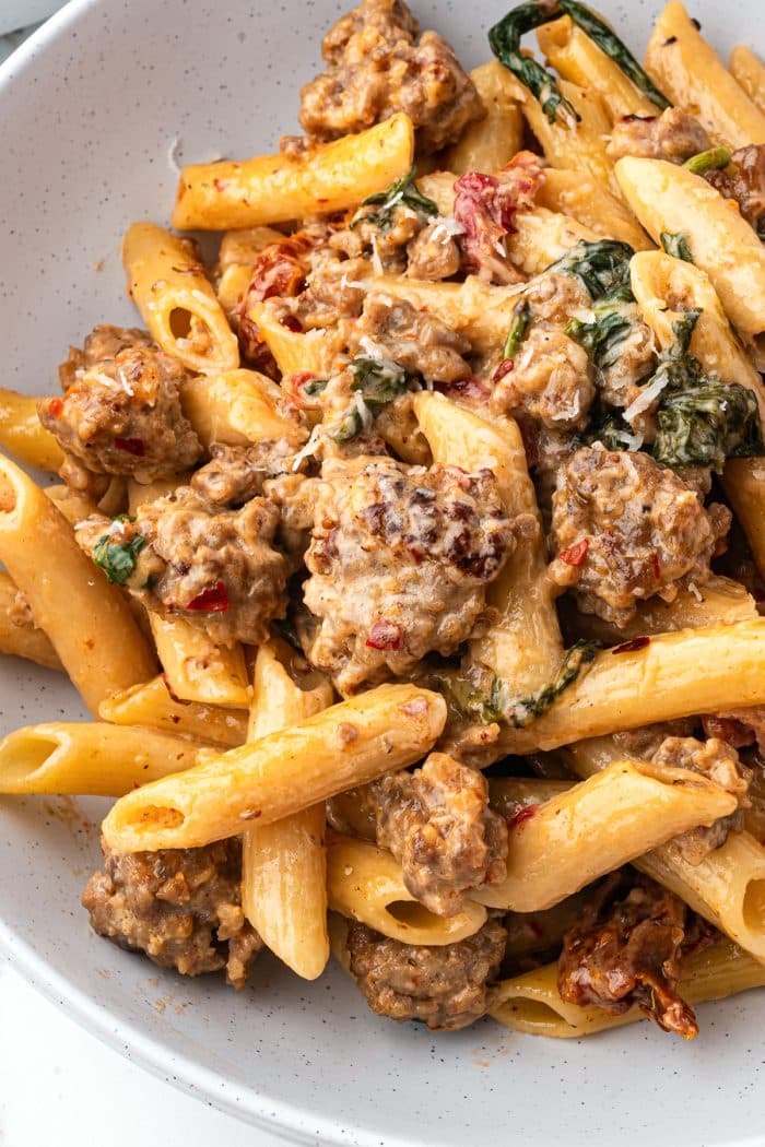 Creamy sausage pasta served in a white speckled bowl with a wooden spoon.