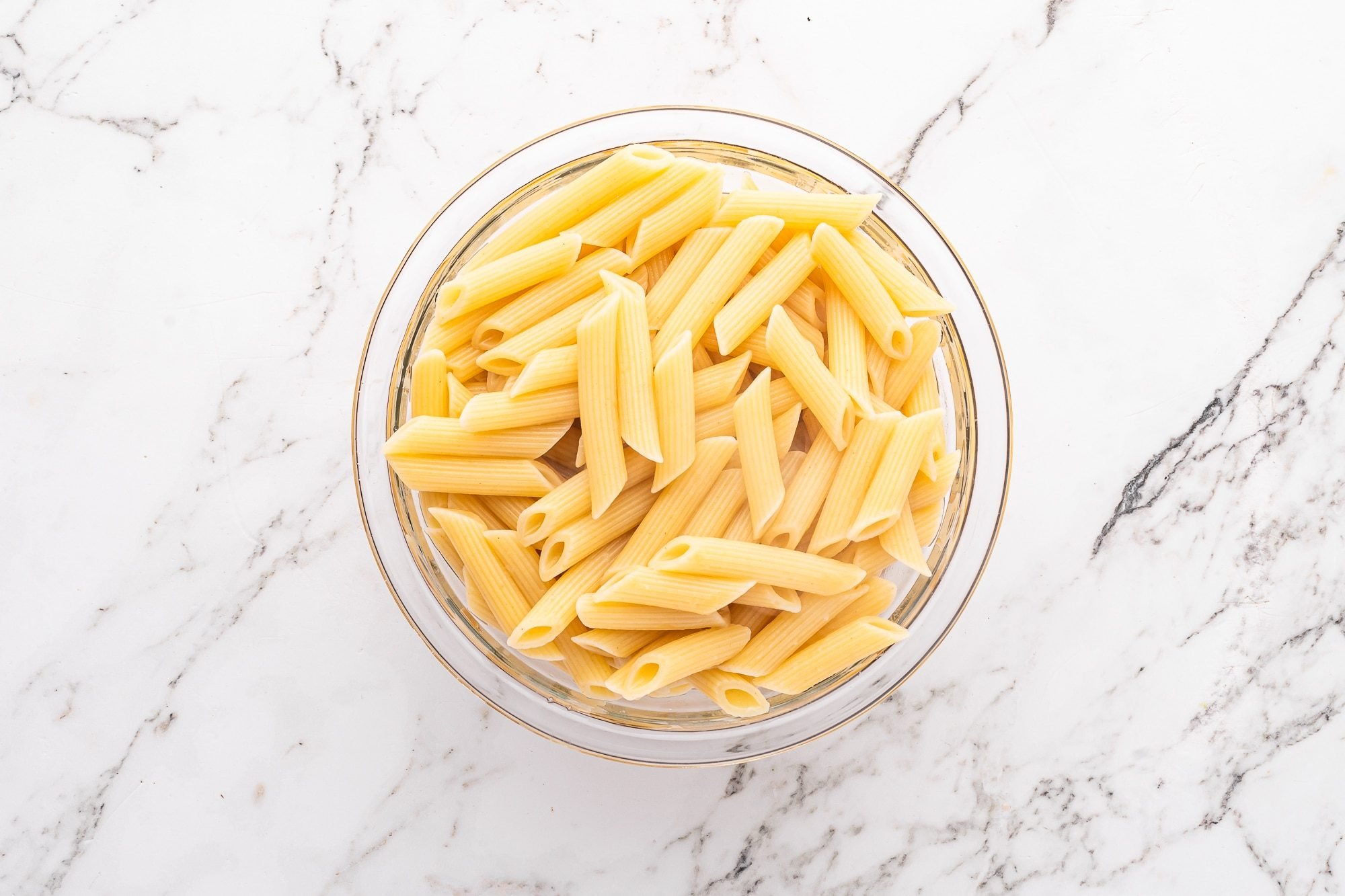 Overhead view of a glass bowl filled with cooked penne pasta on a white marble surface.