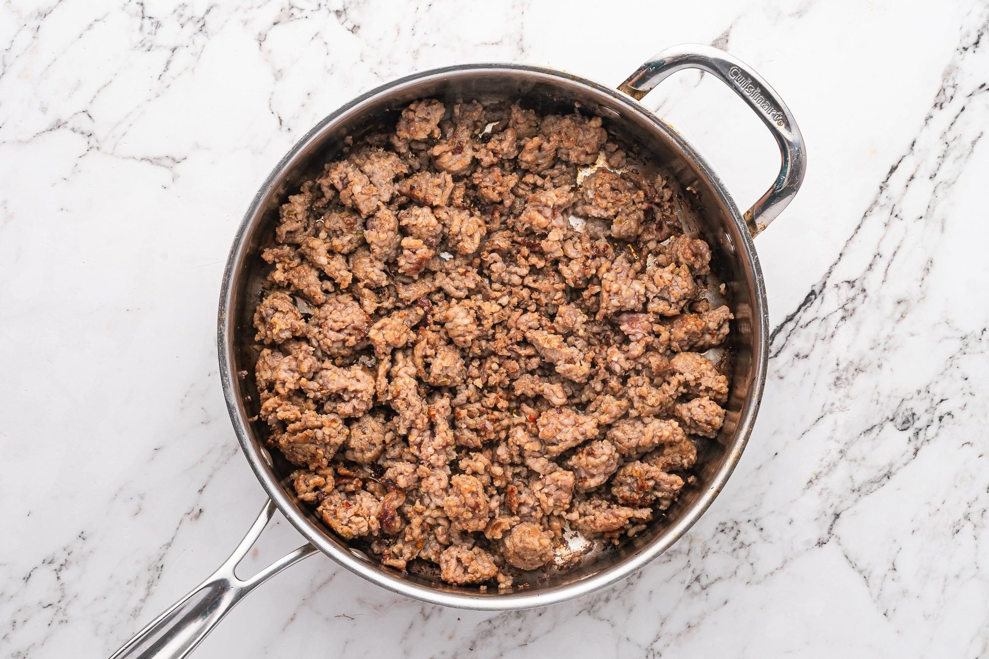 Crumbled Italian sausage browned in a stainless steel skillet on a marble surface.
