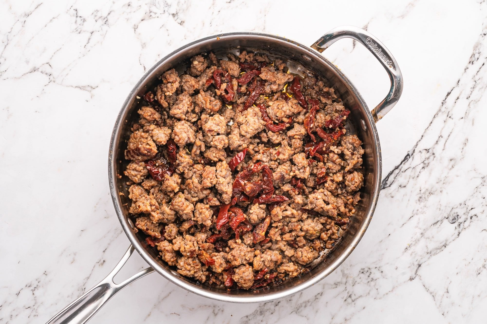 Crumbled sausage cooked with sun-dried tomatoes in a stainless steel skillet.