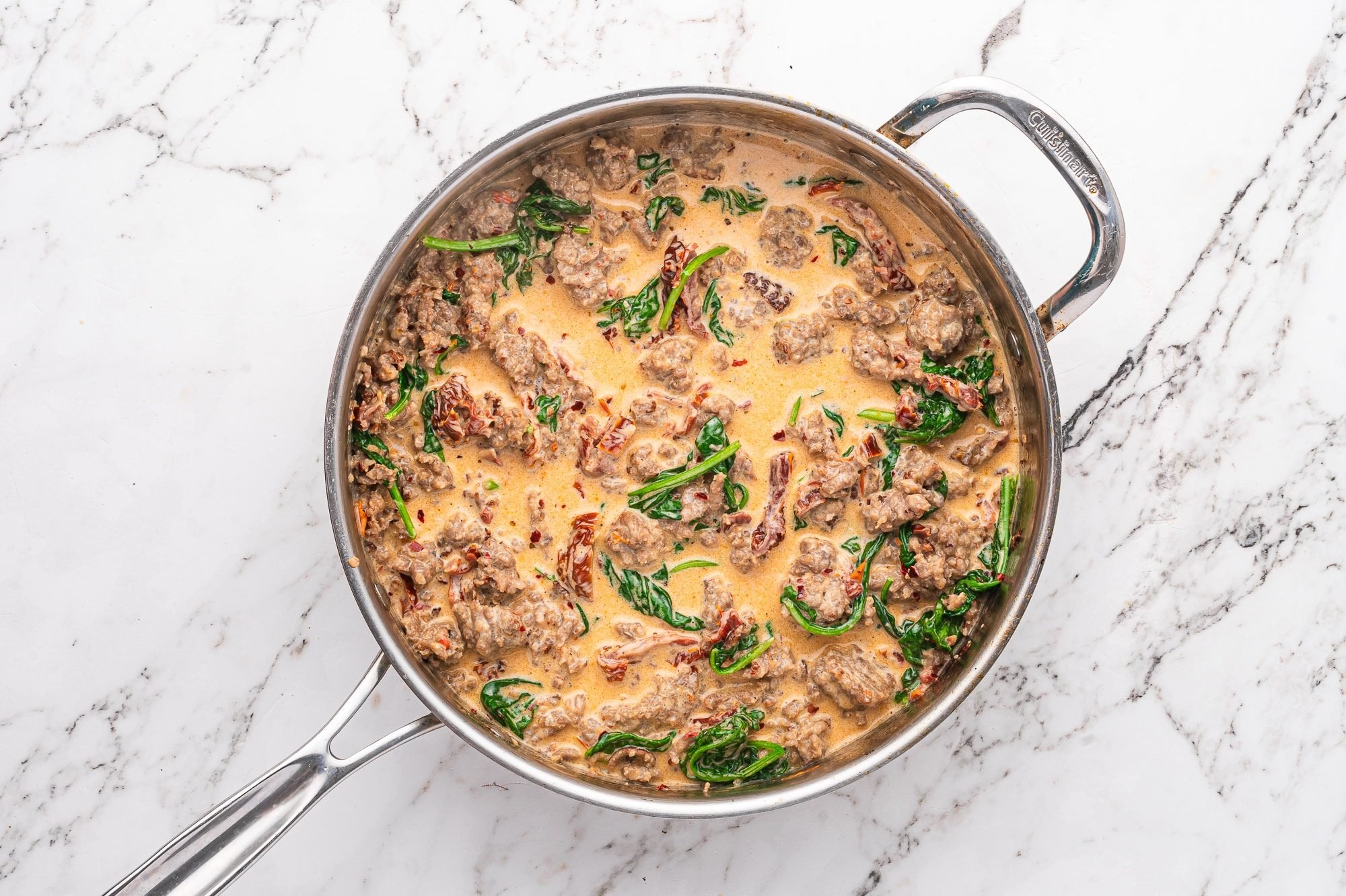 Fresh spinach leaves wilting into a skillet of sausage, sun-dried tomatoes, and cream sauce.
