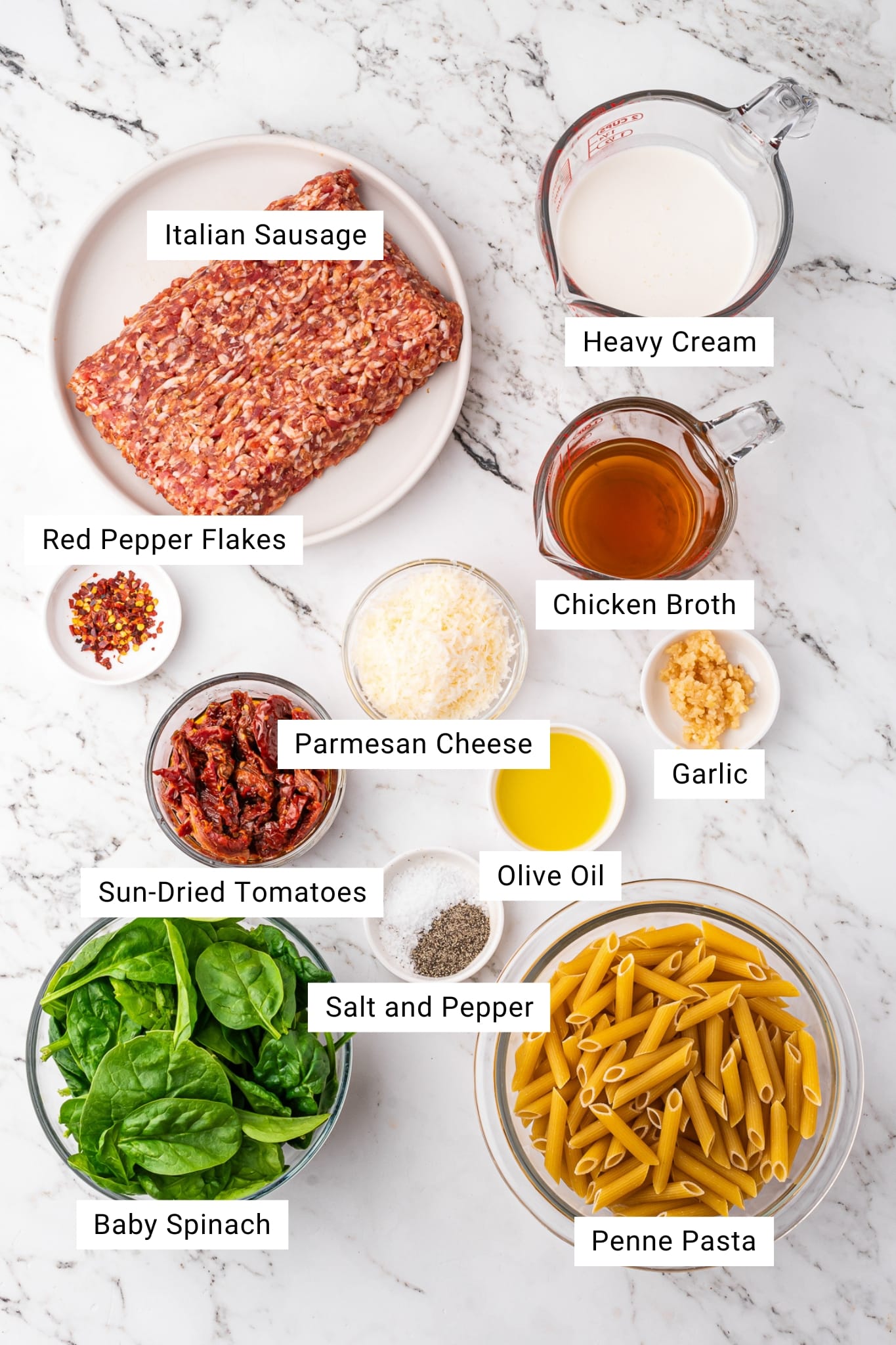 Overhead view of labeled ingredients for creamy sausage pasta arranged on a white marble surface, including raw Italian sausage, dry penne pasta, baby spinach, sun-dried tomatoes, grated Parmesan cheese, garlic, olive oil, chicken broth, heavy cream, red pepper flakes, and small bowls of salt and black pepper.