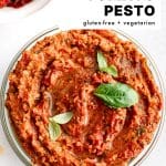 A close-up, overhead shot of sun-dried tomato pesto in a glass bowl, topped with fresh basil leaves. The bold overlay text reads "SUN-DRIED TOMATO PESTO, gluten-free + vegetarian," with "The Forked Spoon" noted at the bottom.