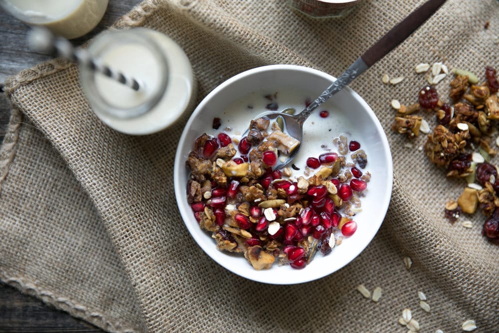 Overhead image of a table with a canister of spiced granola, bowl of pomegranate arils, cup of milk, and bowl filled with a spoon and a serving of crunchy fall spiced granola.