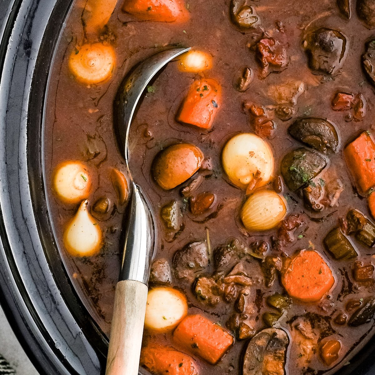 Slow cooker filled with slowly cooked beef bourguignon with carrots, pearl onions, celery, and tender chunks of beef.