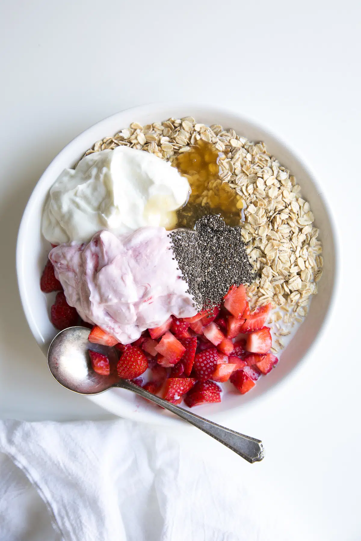 Large white bowl filled with plain yogurt, strawberry yogurt, rolled oats, honey, chia seeds, milk and chopped strawberries.